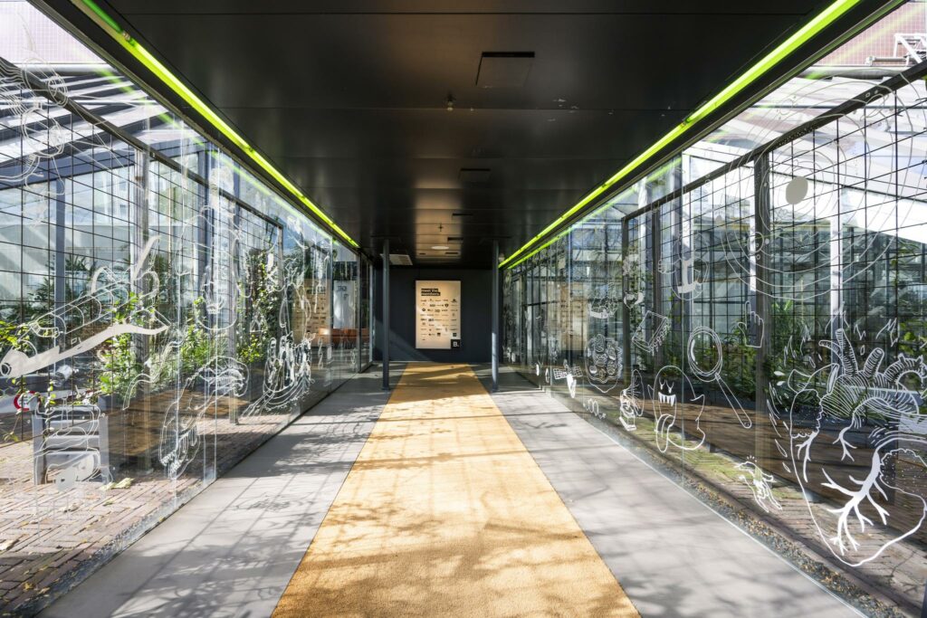 Glazen gang aan de Johan Huizingalaan met tekeningen op de ramen en een geel tapijt op de vloer.